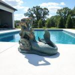 Life-size bronze mermaid statue installed by a luxury outdoor swimming pool in a coastal garden estate.